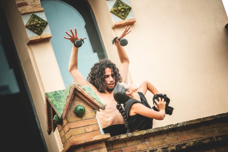 Hombre sin camisa con pelo largo y los brazos levantados bailando con una chica en un balcón de una casa